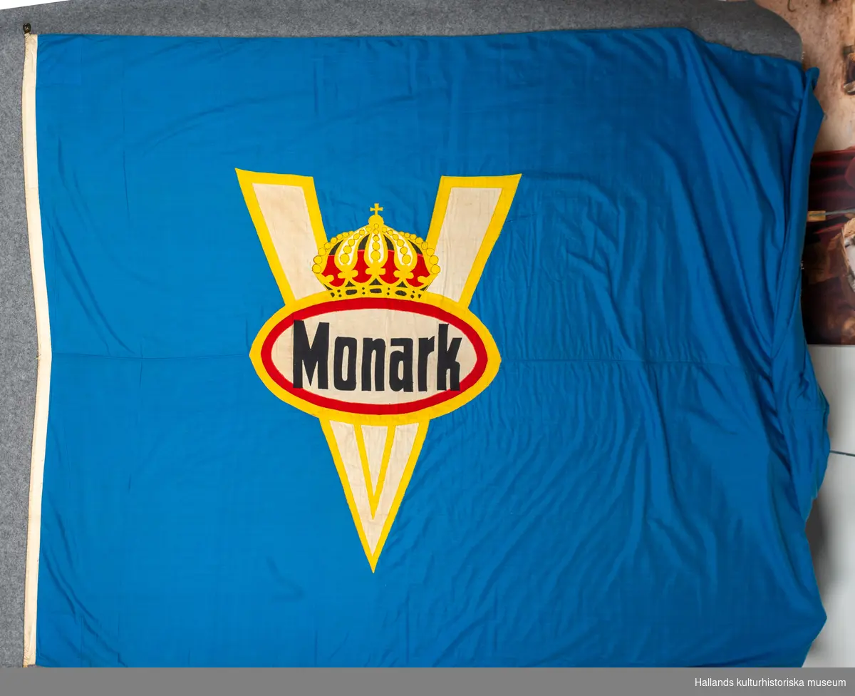Flagga med Monarks emblem, dubbelsidigt applicerad i vitt, gult, rött och svart mot blå botten. Syntet (?)