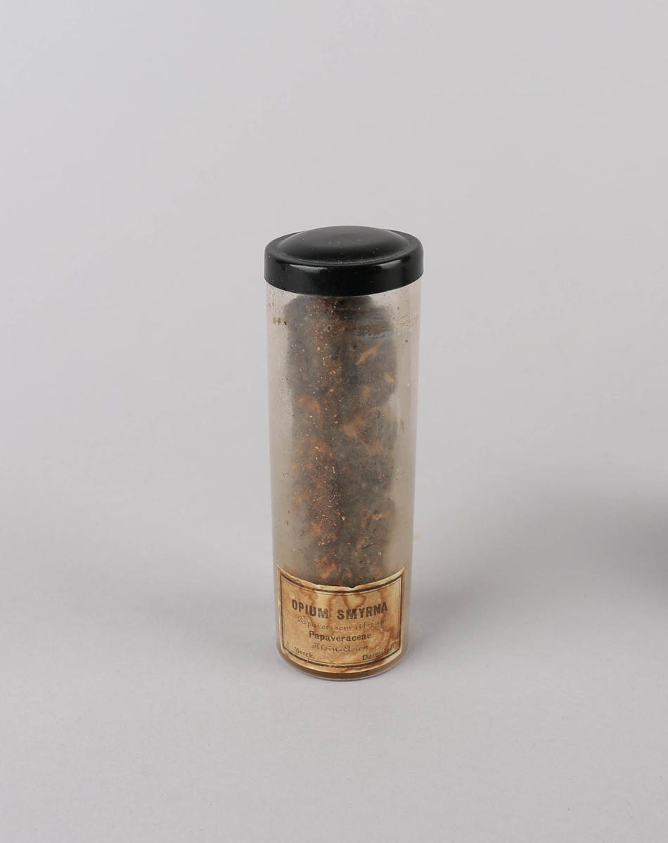 Tørket Opium smyrna (opium) oppbevart i glassylinder av klart glass med lokk. Drogen har brun farge. Beholderen har gulnet etikett med sort skrift og ramme.