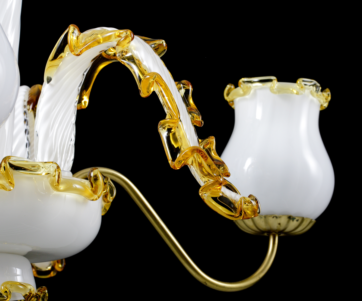 Ljuskrona i venetiansk stil med metallmittstång, varpå ett antal vita och bernstensfärgade glasdetaljer trätts, tillsammans med två "koppar" med fästen för ljusarmar och garnityrer. Ljusarmar i mässing, men i övrigt är alla synliga delar i glas.
