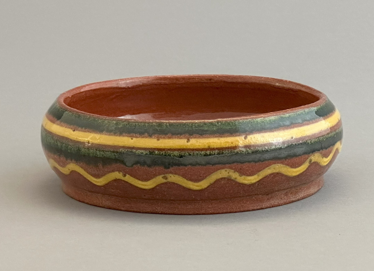 Rödbrun skål av lergods med gul och grön dekor på utsida.