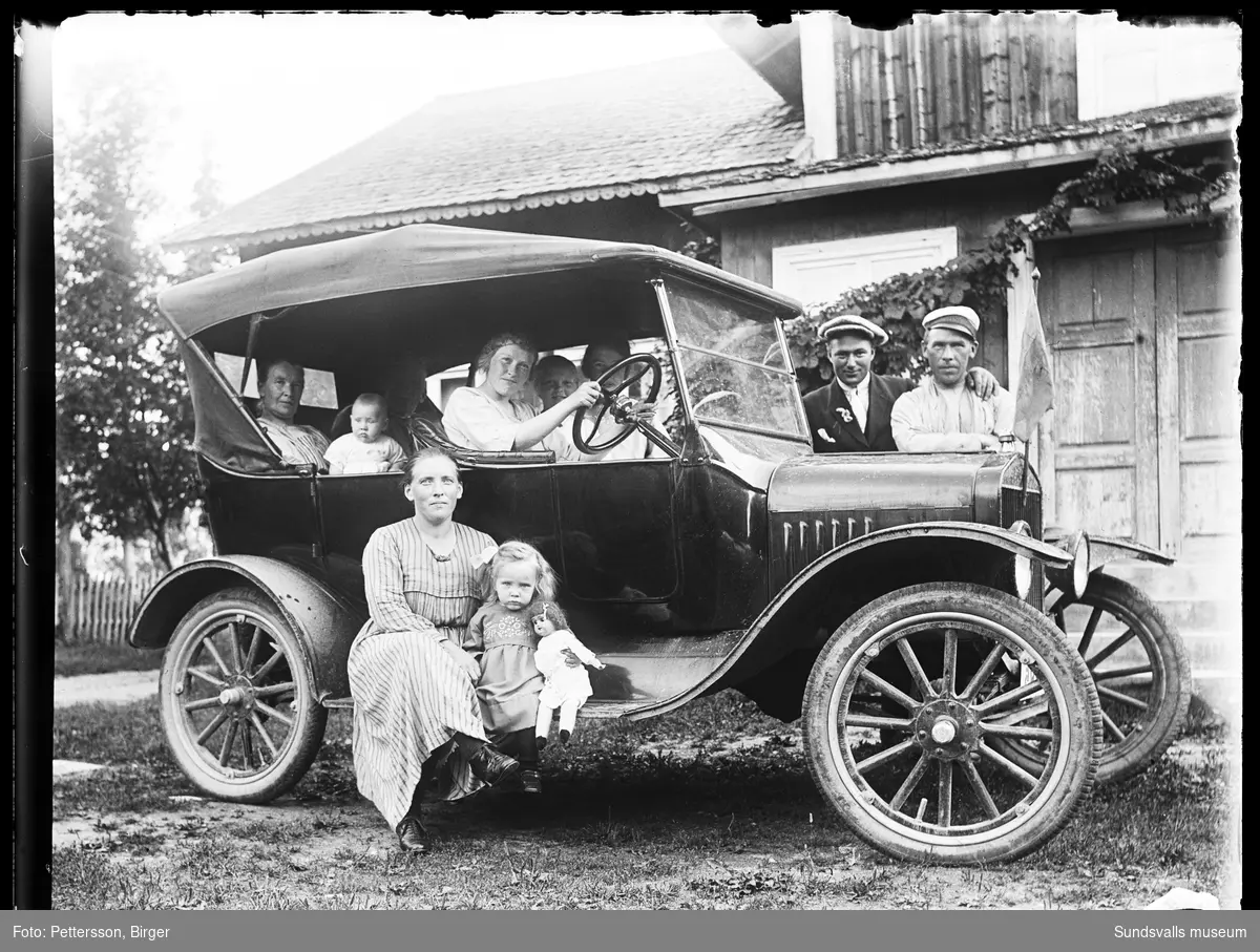 En grupp vuxna och barn i olika åldrar i och invid en ålderdomlig bil som står framför en bostadsbyggnad. Samma personer som SuM-foto035232. Bilen ser ut att vara samma som SuM-foto035231, en T-Ford från ca 1923-25 och med registreringsnummer Y285.