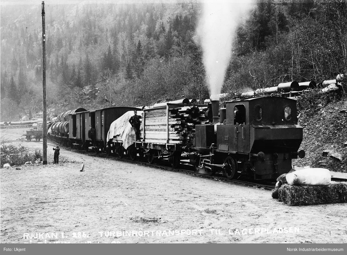 Tog transport av turbinrør til lagerplass på Såheim. Lokomotivet Odin og lukkede godstvogner type Litra G3 og plattformvogner. På jernbanespor på siden står en kassevogn. Ansatte på jernbanen i lokomotivet og vognene. Turbinrør lagret i lia ovenfor toget.