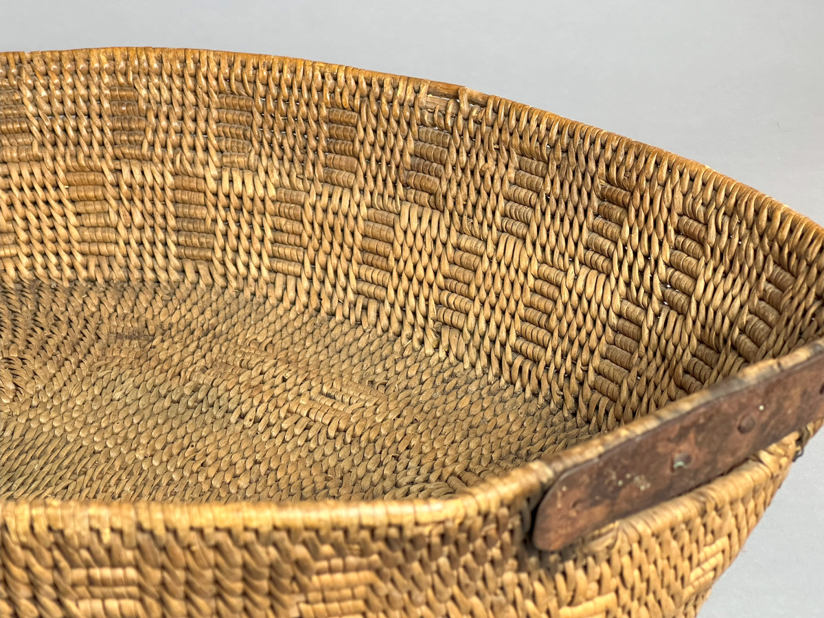 En oval såkorg, tätbunden av rot med mönster. Två handtag, varav det ena är lagat med en metallskena.