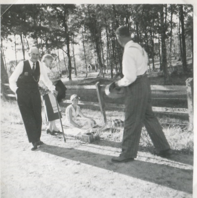 Fyra vuxna personer befinner sig på gårdsplanen vid Backen eller Högen 1960-tal. Från vänster: 1. Okänd äldre man med käpp. 2. Ester Pettersson (med förkläde, 1894 - 1988). 3. Okänd sittande kvinna. 4. Gående man.