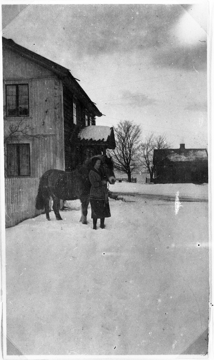 Marie Sandberg gm Karl Larsen holder en hest foran hovedbygningen i Ekeren nordre.  Vi ser litt av gavlveggen mot syd som er bordsydd og malt og østveggen som vender mot gardsplassen.  I bakgrunnen til høyre ser vi en gammel størhusbygning.