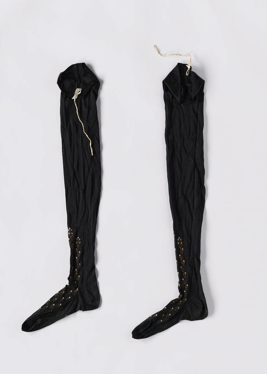 Ett par strikkede svarte nylonstrømper. Foten er formsydd. Det er broderinger i blomstermønster fra oversiden av foten og oppover leggen. Det er bånd i kanten, og hull i brettekanten for å tre inn bånd.