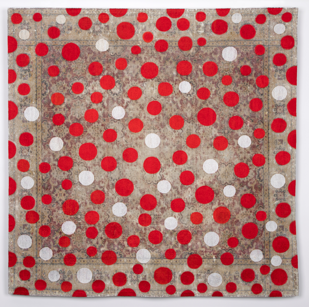 Kvadratisk veggtekstil av fløyel med trykt mønster som minner om persisk teppe. Stoffet har et tynt lag med hvit akrylmaling. Håndbroderte sirkler i hvit og rød bomullstråd.