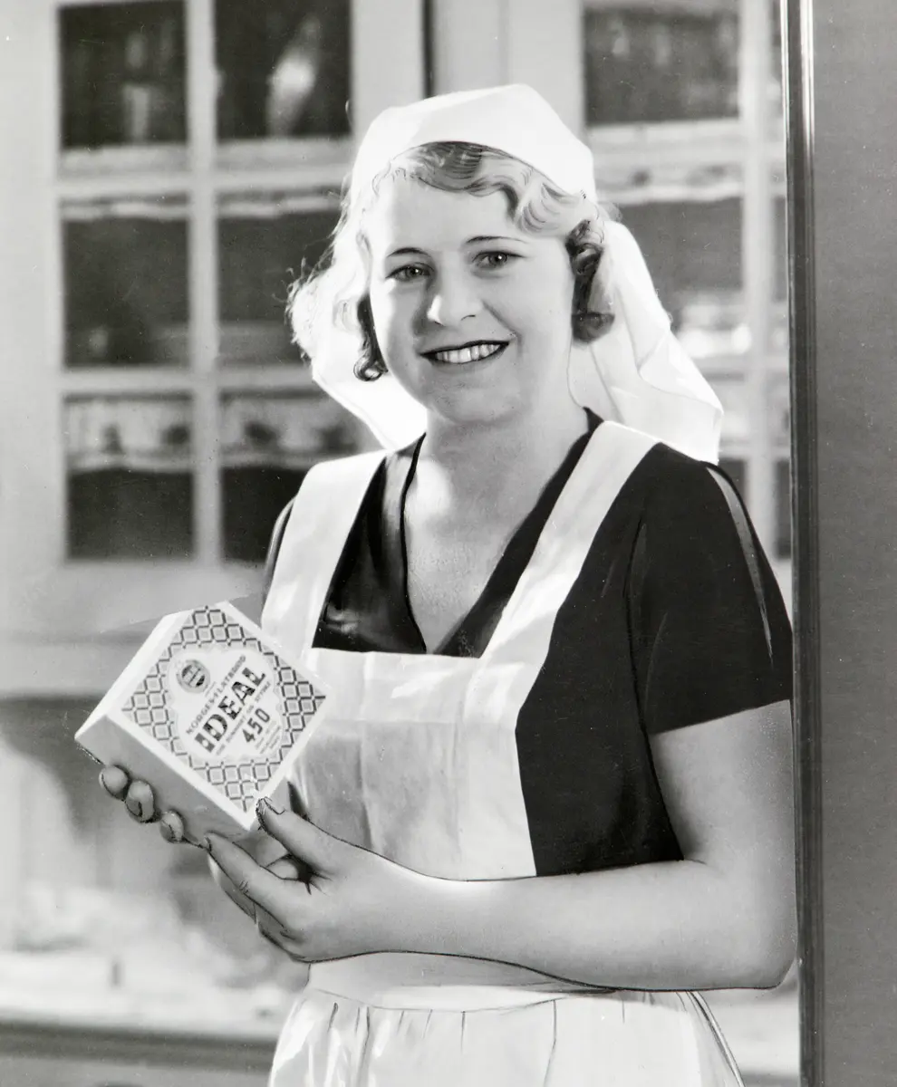 Svart-hvitt foto av blond dame med skaut og hvitt forkle som står med en pakke Ideal-flatbrød i hendene.