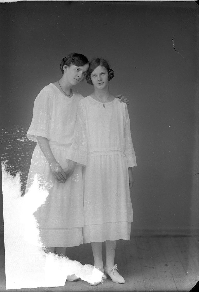 Två unga kvinnor i ljusa tunna klänningar stående tätt intill varandra.