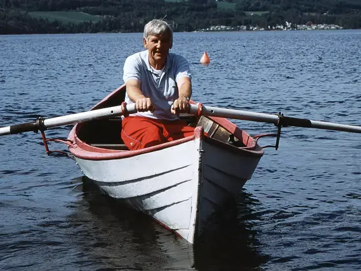 En mann sitter i en hvit og rød trebåt og ror på vannet. 