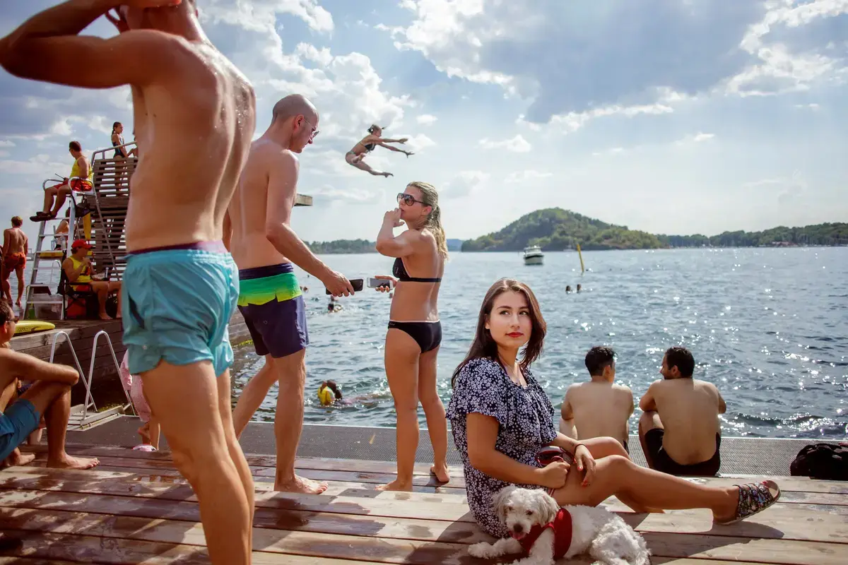 Bilde av mange mennesker delvis på en brygge og i vannet. En kvinne sitter i midten med en liten hvit hund ved sin side.