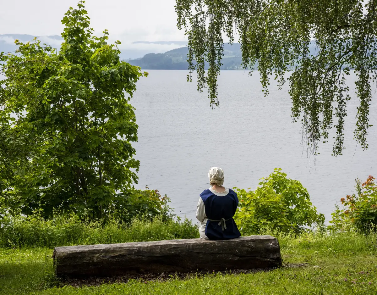 Ei dame kledd i enkle middelalderklær og med hodeplagg, sitter på en benk og skuer ut over Mjøsa.