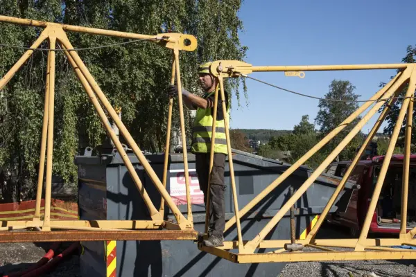 En mann med hjem og refleksvest kobler sammen to gule stålelementer til en byggekran.