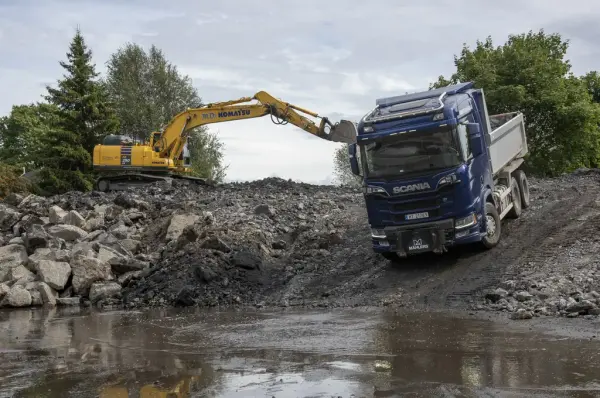 En gul gravemaskin står oppe på en haug med grabben strekt ut, en blå Volvo lastebil med grått plan står på skrå ned mot en dam med sølevann.