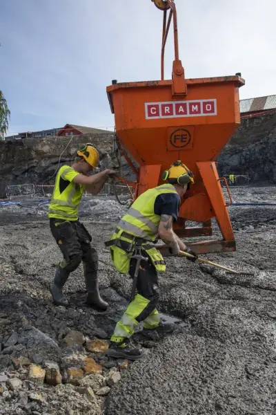 En mann med hjelm og gul signalvest styrer en stor, oransje betongkjele mens en annen mann i like klær glatter betongen utover med rake.