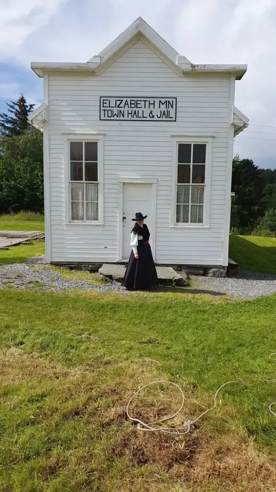 Kvinne i drakt som passer Rådhuset fra 1880-tallet. Hvittmalt amerikansk rådhus i med høyrest falsk fasade