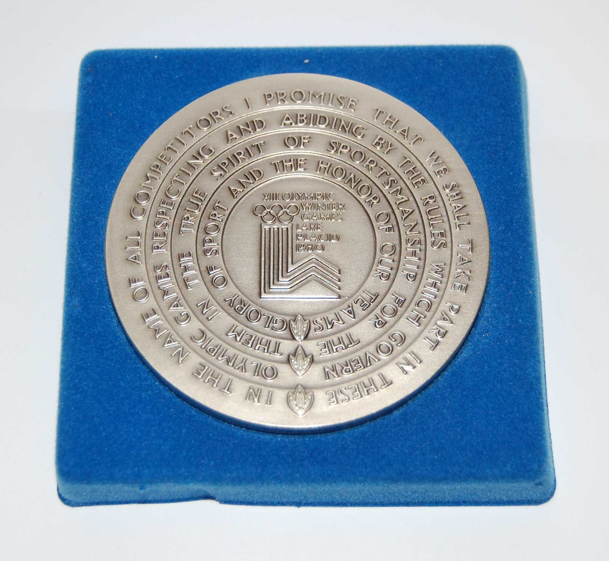 Minnemedalje med emblemet for de olympiske vinterleker i Lake Placid i 1980 omringet av teksten den olympiske ed. På medaljen er det også motiv av ulike idrettsutøvere som driver forskjellige grener. 
Medaljen ligger i et blått etui.

Deltagermedalje. Ligger i turkis eske.