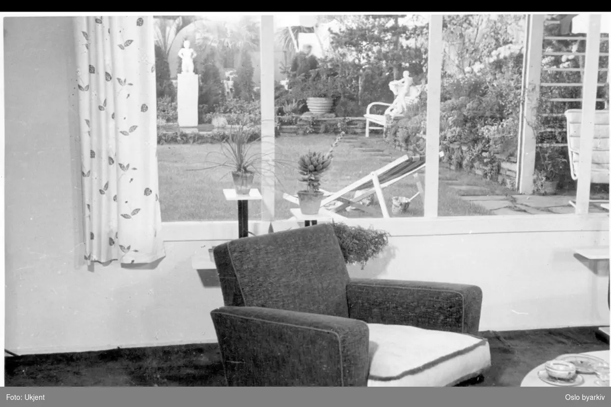Utstilling i Roald Amundsens gate, "Blomstene i hjemmet". Innsyn fra husets dagligstue mot blomsterutstillingens have. Plen, havemøbler, (fluktstol), basseng med skulptur, blomsterbed, trær og prydvekster. Albumtittel: "Utstillinger".