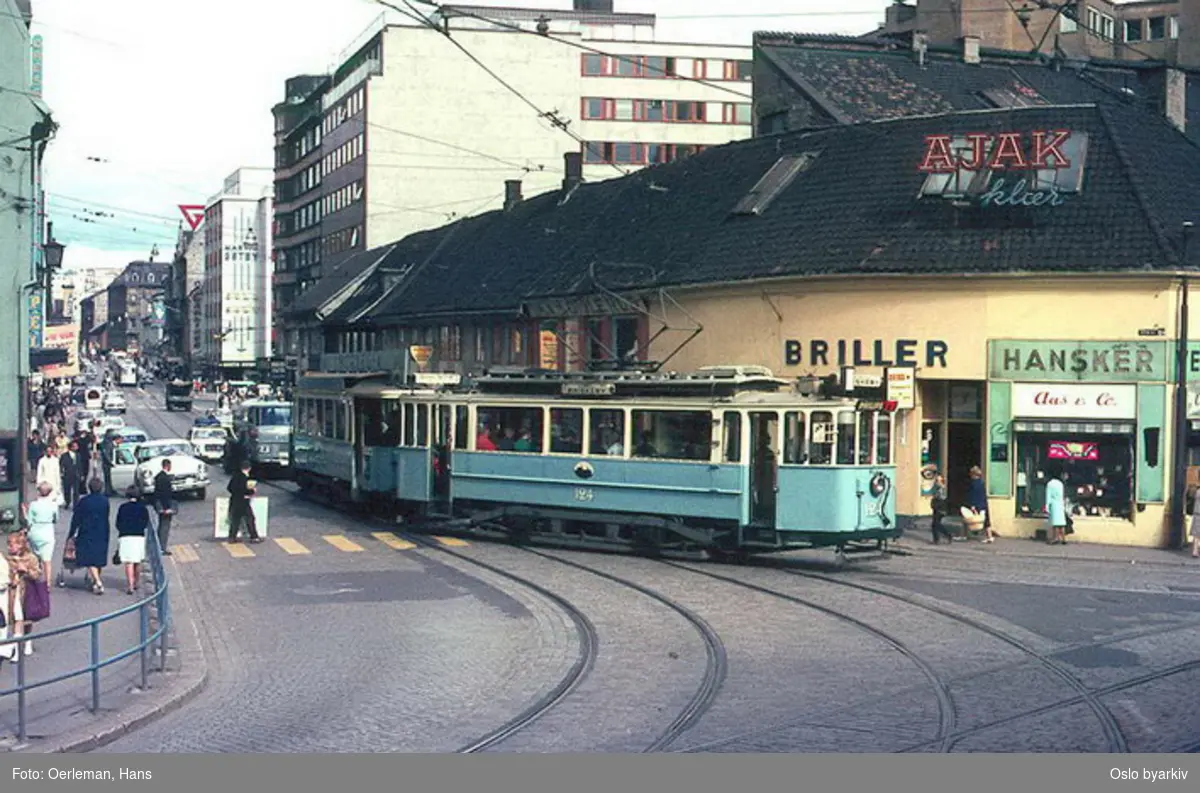 Oslo Sporveier. Trikk motorvogn 124 type HaWa linje 1 svinger ut av Storgata ved Kirkeristen. Brille- og hanskebutikk, butikkskilt.