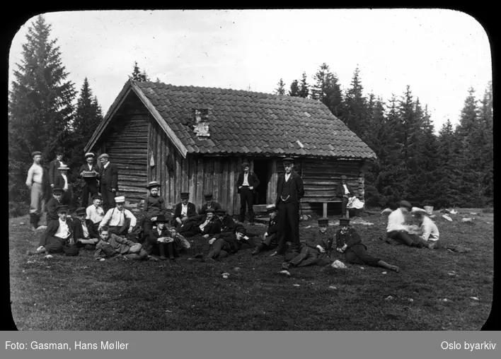 En gruppe menn og ungdommer poserer for fotografen på vollen foran stølshuset på Vensåsseter i Bærumsmarka, , 5. juli 1903