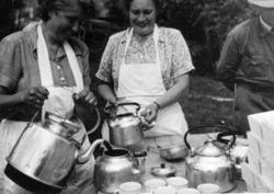 To damer med forklær skjenker kaffe. Kaffekjeller, kopper, ø