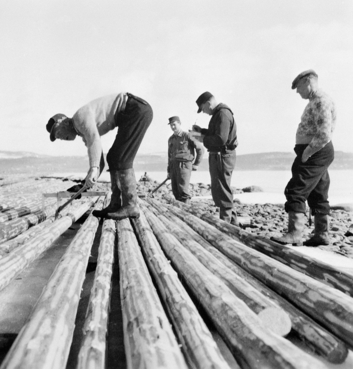 Gruppe menn klaver (måler), stikker (fløyholder) med stikkbok, to påslåere. Tømmermåling på stranda ved Mjøsa på Grefsheim, Nes, Hedmark.s.