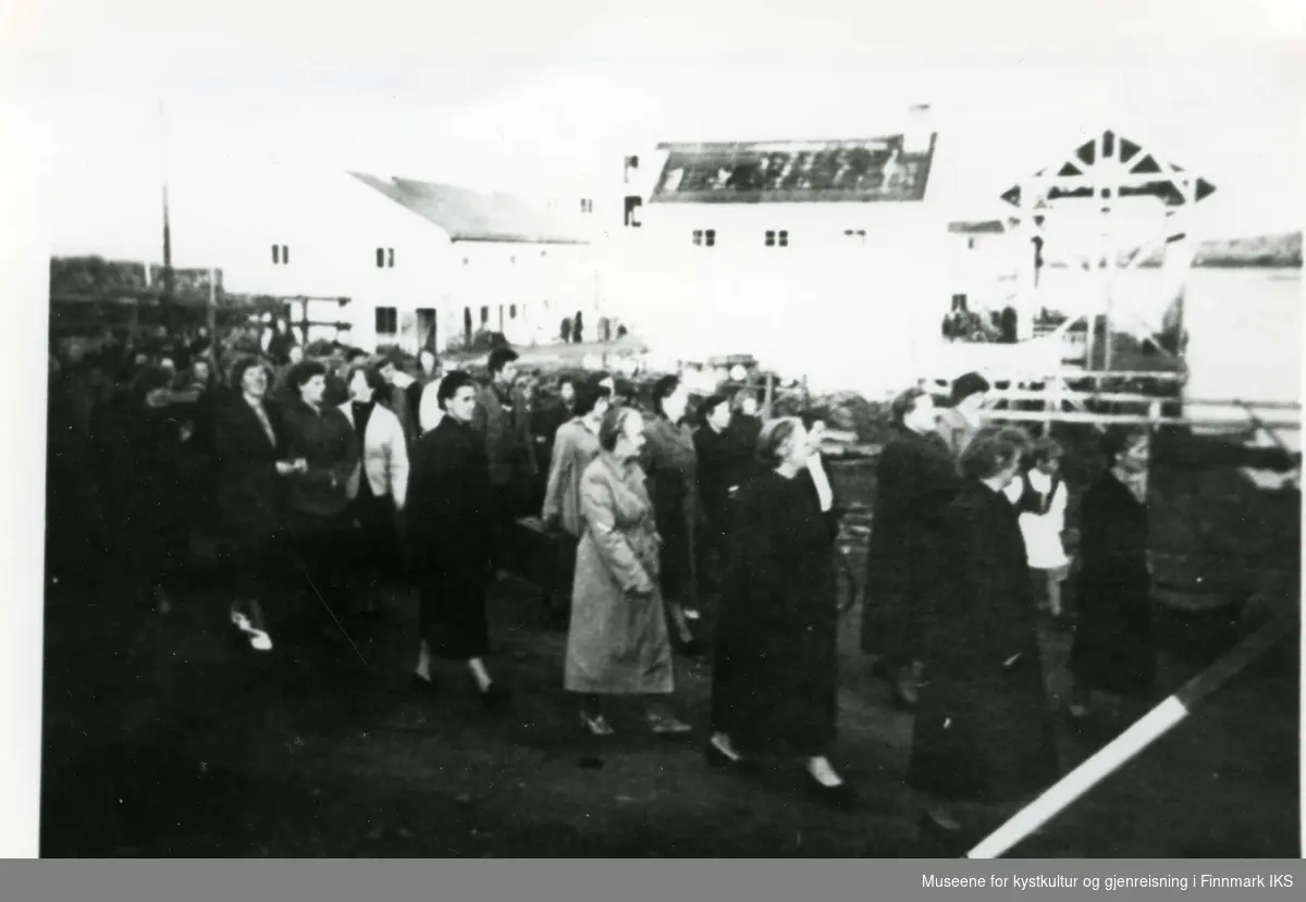 Bildet er tatt på Nesset i Gamvik. Menneskene på bildet er deltagere på Husmorlagsstevnet. Disse er kommet med Hurtigruten. Dampskipskaien ligger bak bygningene i bakgrunnen. (Produksjonslaget.)  1957/58