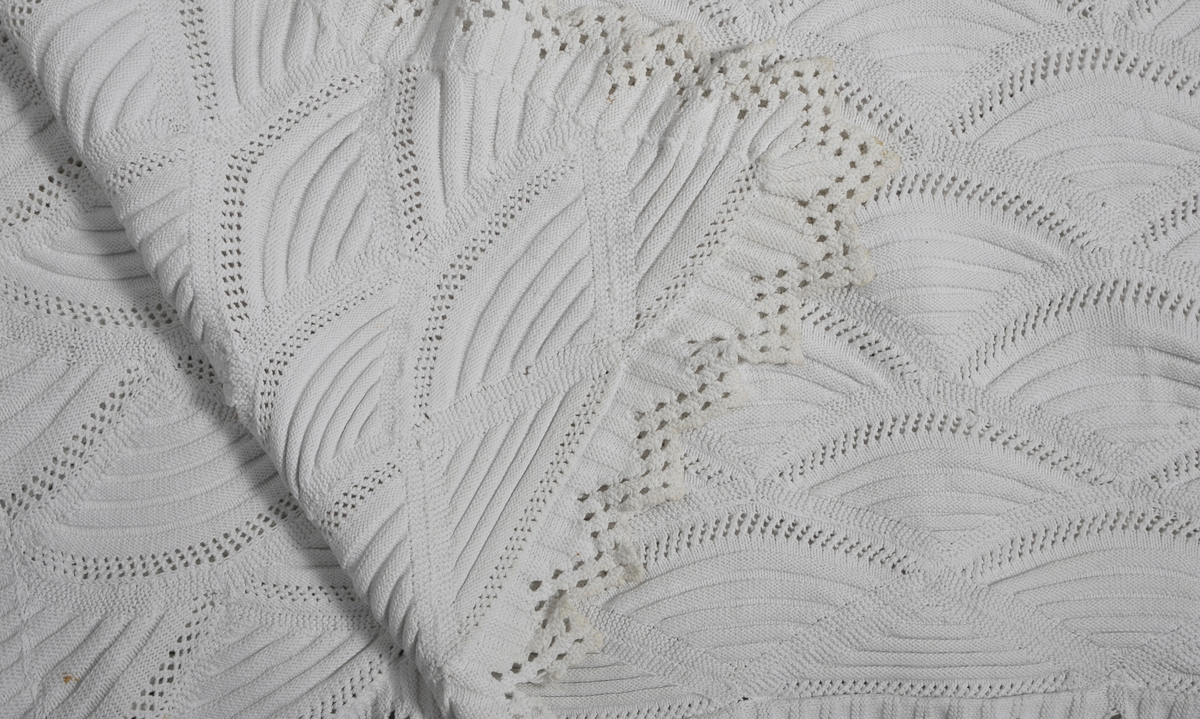 Strikket sengeteppe med mønstereffekt i vifteform. Rett og vrangmasker utgjør mønster. Rundt teppet er det påsydd en strikket kant.