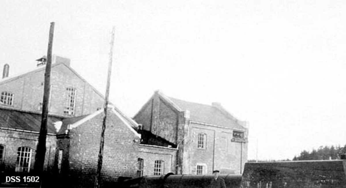 Namdal tresliperi.  Denne treforedlingsbedriften ble etablert etter initiativ fra Odd Soot-Ryen (1865-1928) og Karl Kristian Greiff (1854-1919) i 1909 på et sted der det tidligere hadde vært sagbruksvirksomhet under navnet «Løfsnæs Brug».  Fotografiet viser Industribygninger i upusset teglstein med vinduer i støpejernsrammer.  I forgrunnen en turbinledning.  Mannen som står foran ledningen skal være sliperimesteren.