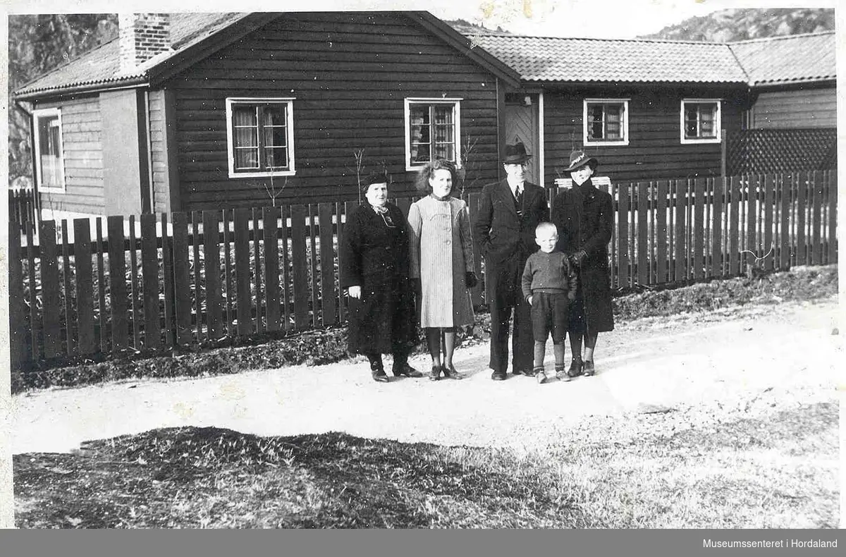 Folk i finklær, hus, tregjerde, veg, litt natur.