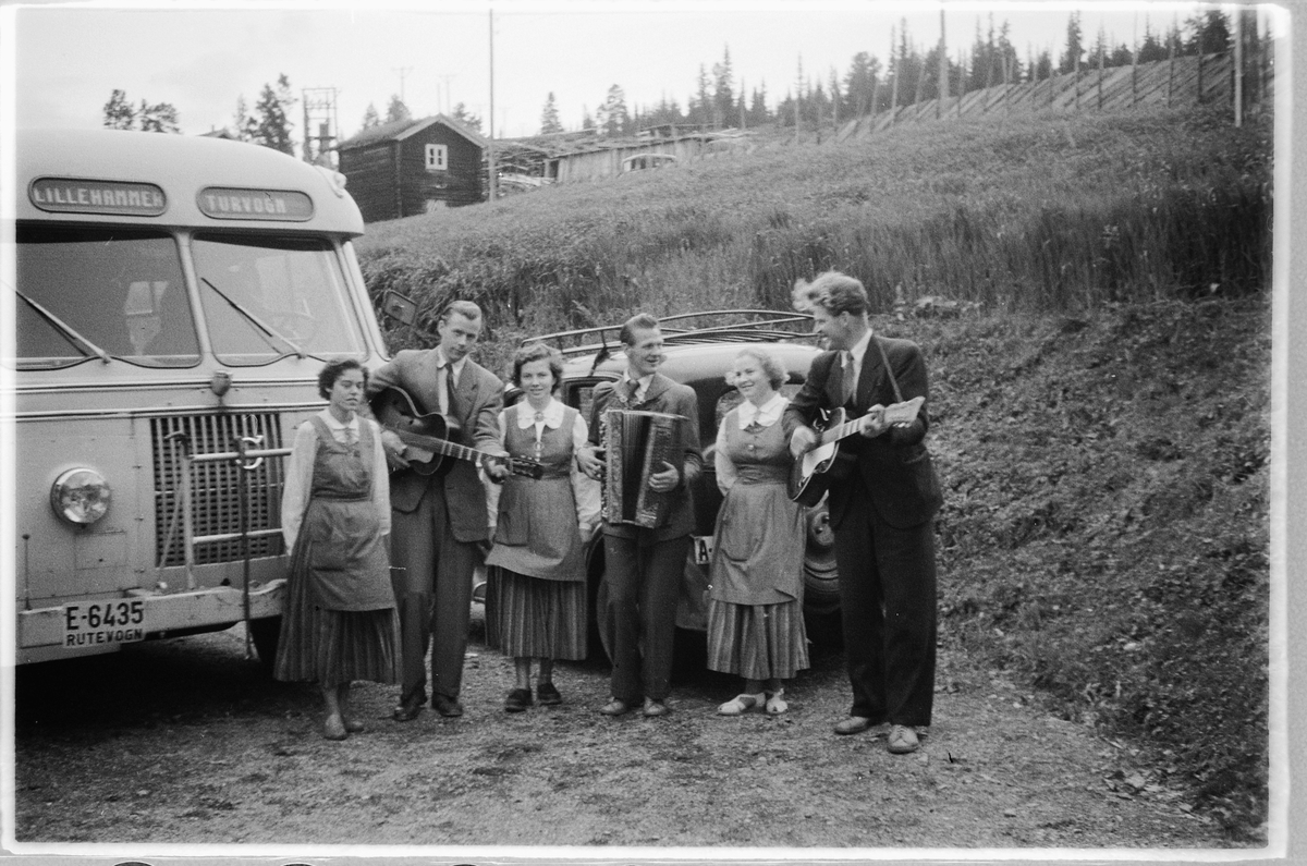 Tre menn spiller gitar og trekkspill, mens tre kvinner lytter ved en buss med skilt Lillehammer turvogn. T. h. Ulf Sæhli på gitar. Volvo buss E-6435.