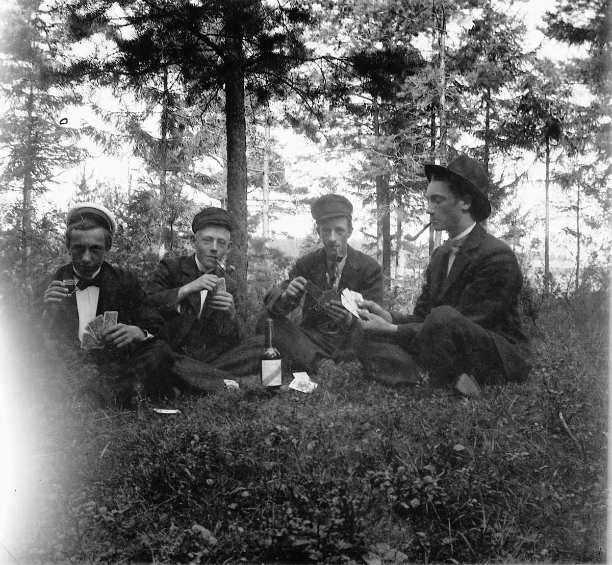 Eksteriør, skog. Fire unge menn drikker, røyker og spiller kort. 