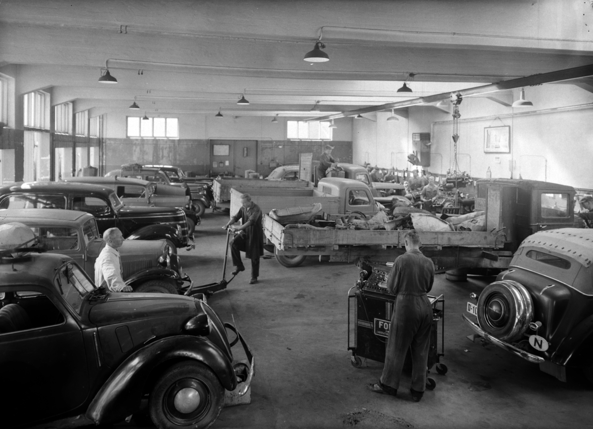 A/S Opplandske Automobilforretning, biler, arbeidsfolk, div. Motiv, Enggt. 34, Hamar. Bilverksted. 
Fra venstre side, nærmest Fiat 1100 ca. 1936-39, deretter ukjent, Ford V8 standard varebil <delivery sedan). Til høyre bakparten på en Opel Super Six ca. 1936-38. 