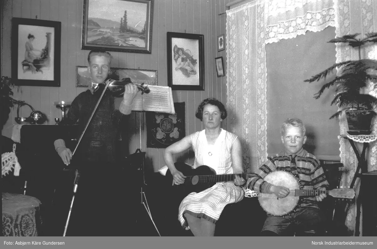 To gutter og en dame spiller på instrumenter i stua: filolin, banjo og gitar. Blomster, møbler og gardiner avbildet  