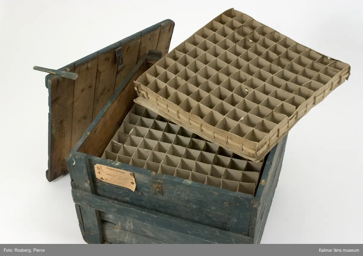 KLM 39806:1. Transportlåda, transportlåda för ägg, trä. Grönmålad. Tvärgående slåar och lås med sprint av trä. Inne i lådan finns insatsfack av papp för ägg, fem lager, 100 ägg i varje lager, lådan kan rymma 500 ägg. En fraktgodslapp utan text sitter fäst på lådan.
