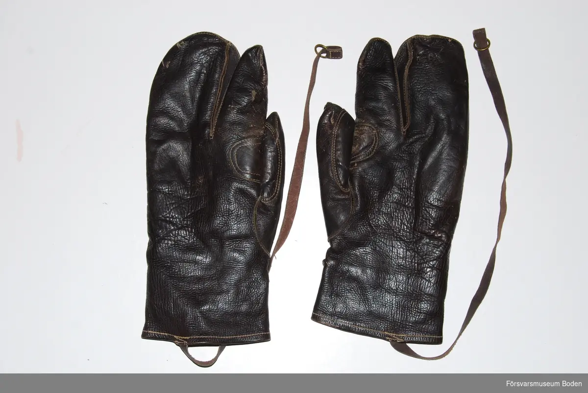 Ett par handskar av trefingersmodell i mörkbrunt skinn. Spännrem runt handleden samt lång rem för fastsättning i fältdräkten. Präglade med tre kronor-symbol. Tillhörande innervantar registrerade under FMB.002605.