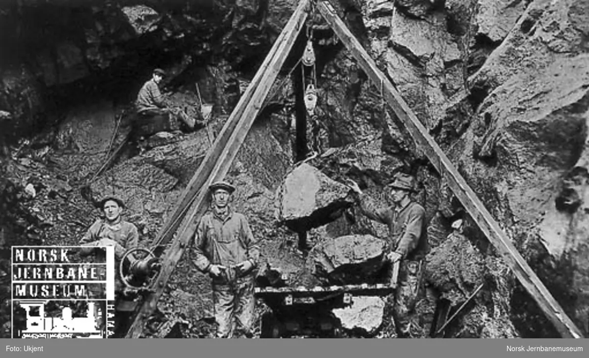 Tunnelanlegg på Sørlandsbanen vest for Kristiansand : fire mann i arbeid, stein løftes opp på en tralle med stubbebryter