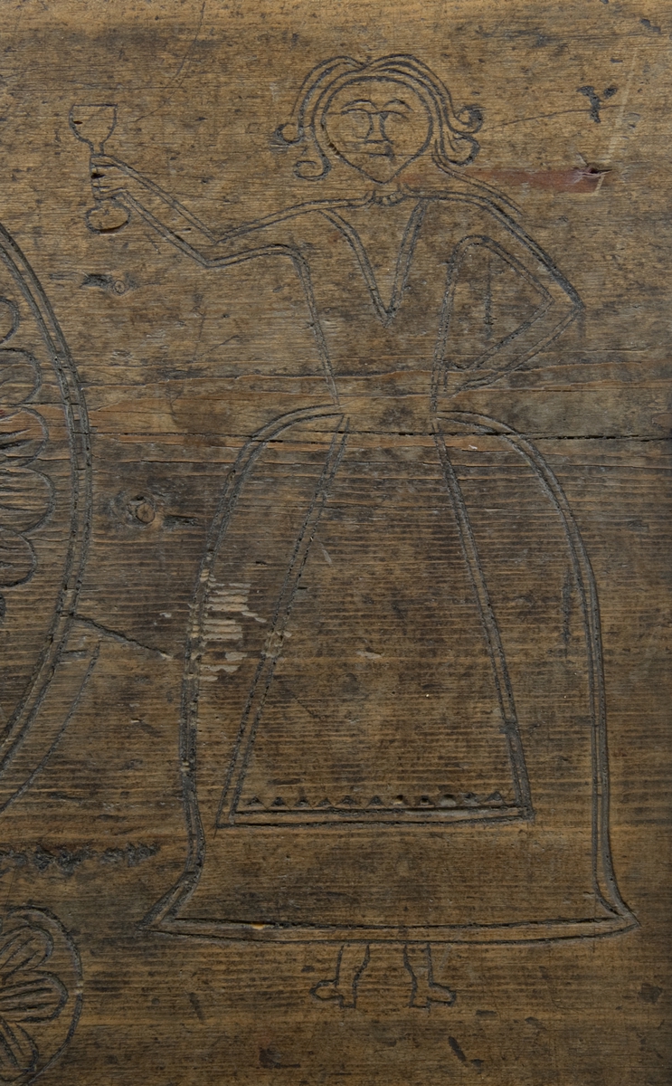 Kiste med flatt lokk og karveskurd på lokk og front. På fronten skåret geometriske figurer og blomster, på lokket skåret en hest, en kvinnefigur, en mannsfigur og en løve.