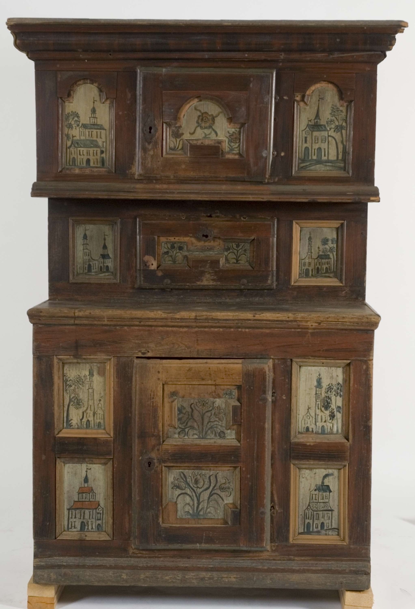 Framskap i en del med en dør nede, en  oppe og en liggende i midten. Dørene har malte blomstermotiver mens de 8 sidefeltene har malte bygninger.