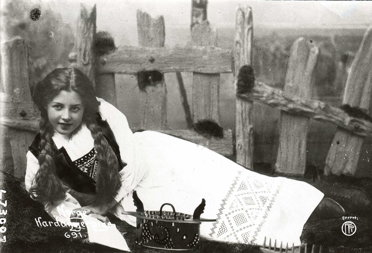 Studiofotografi av en jente som ligger foran gjerde, med tine. Drakt fra Hardanger.