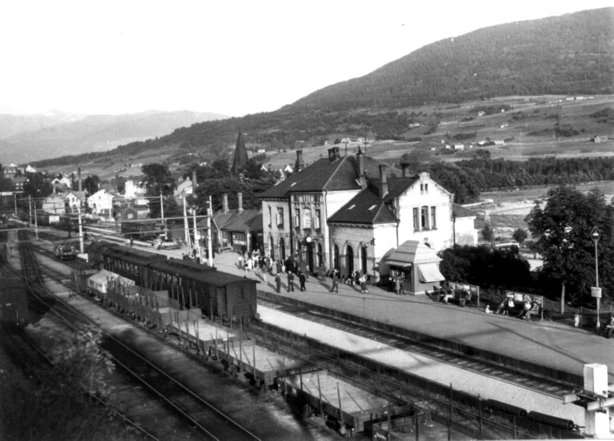 Voss jernbanestasjon 1939. Jernbanevogner og godsvogner på stasjonen. Passajserer på perrongen. Landskap med gårder, jorder og åkrer. Skog og fjell i bakgrunnen.