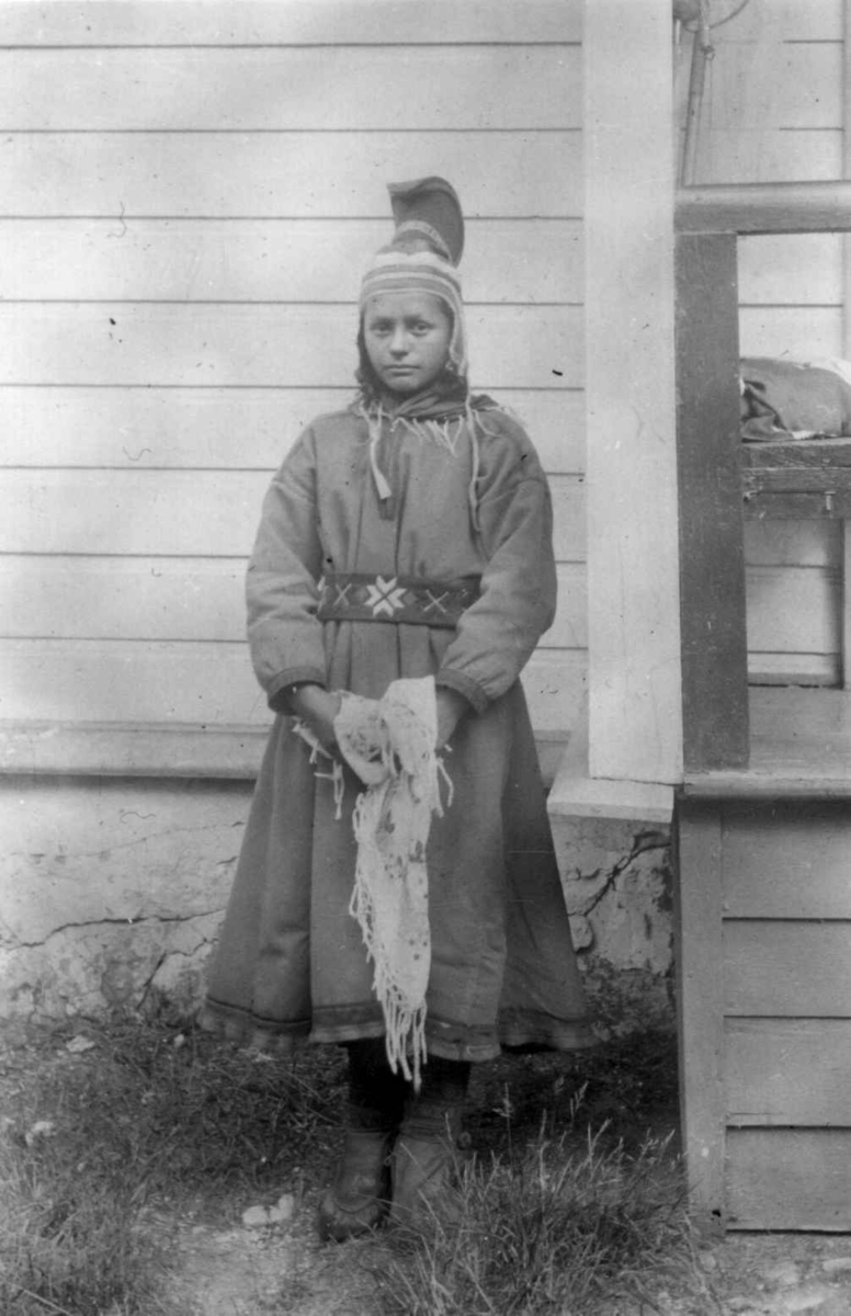 Portrett av samisk pike fra Sandnes, Sør-Varanger, Finnmark. Hun poserer utenfor Solheim, legeboligen til Wessel i Kirkenes, Sør-Varanger, Finnmark. Ukjent år.