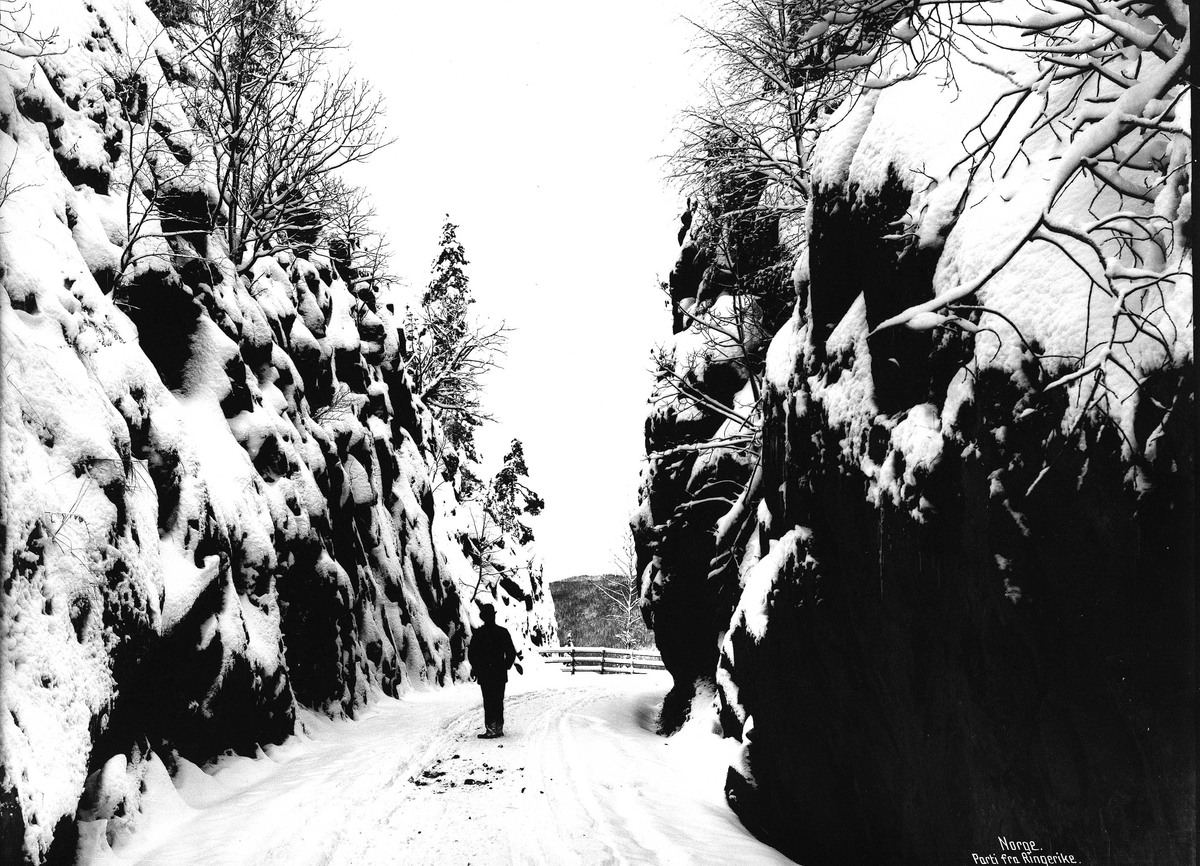 Landskap, veiparti ved Skaret, Hole med fjellvegger på begge sider av veien. En mann står på veien med ryggen til. Del av Den Norske Turistforenings samling landskapsbilder, de fleste tatt av fotografene Brødrene Brunskow.