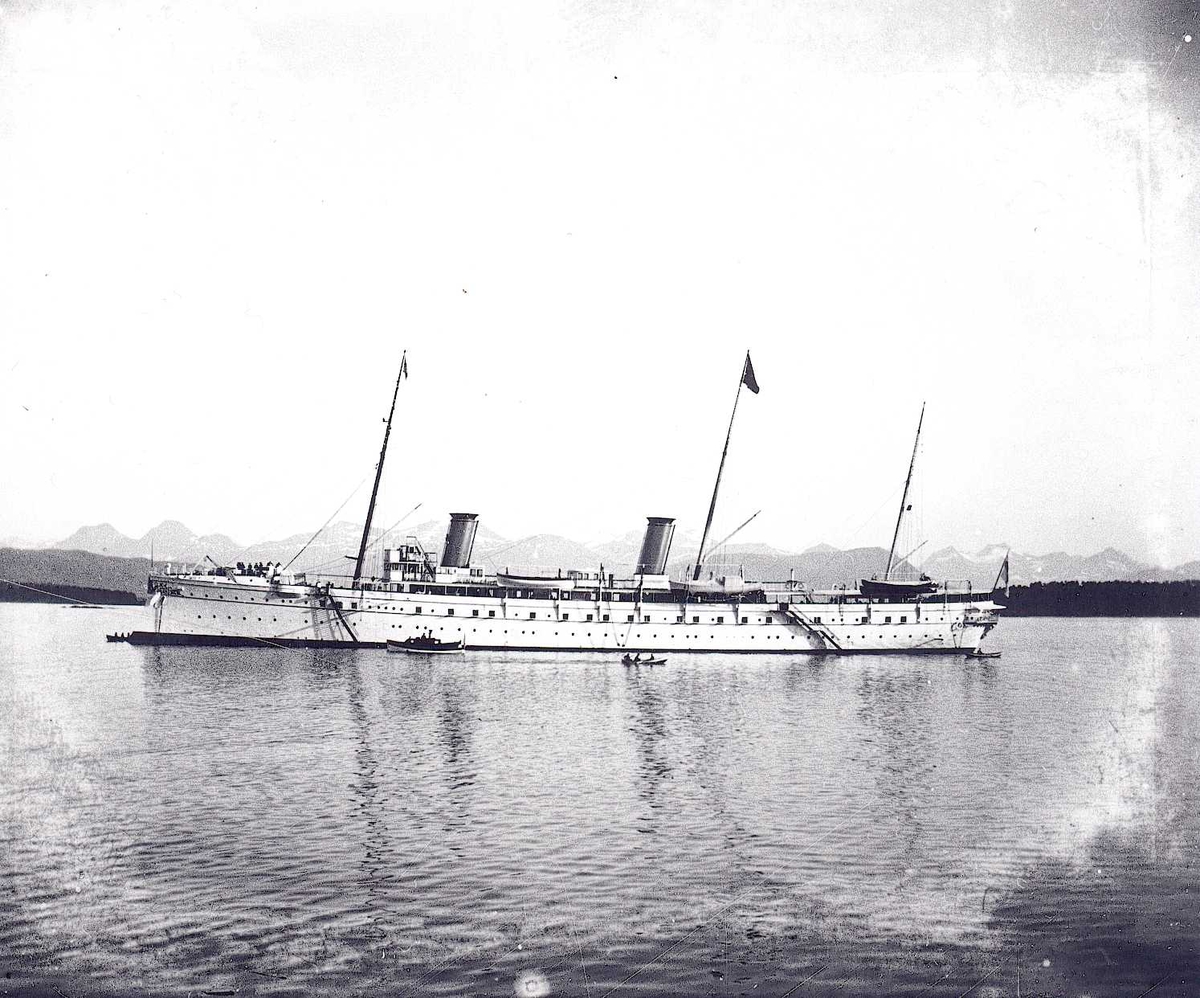 Keiser Wilhelm II' skip S.M.S. Hohenzollern på havna i Molde, under et av sine årlige besøk i perioden 1889-1914. Motorbåt og robåt på siden av skipet. Sunnmørsalpene i bakgrunnen.