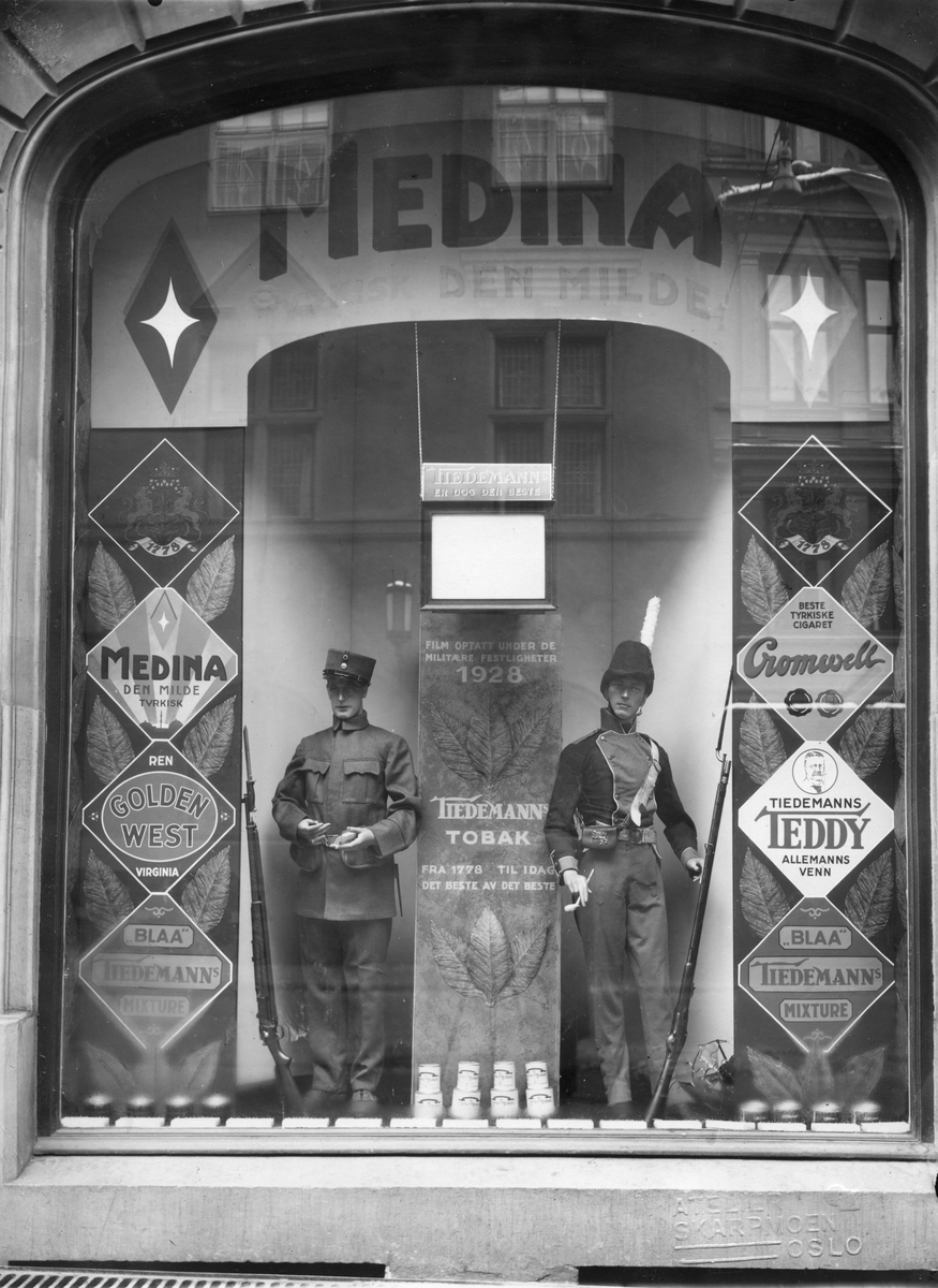 Vindusutstilling med reklame for Tiedemann. To utstillingsdukker med militæruniformer røyker pipe og sigarett. Man viser også en film som er optatt under de militære festligheter 1928.