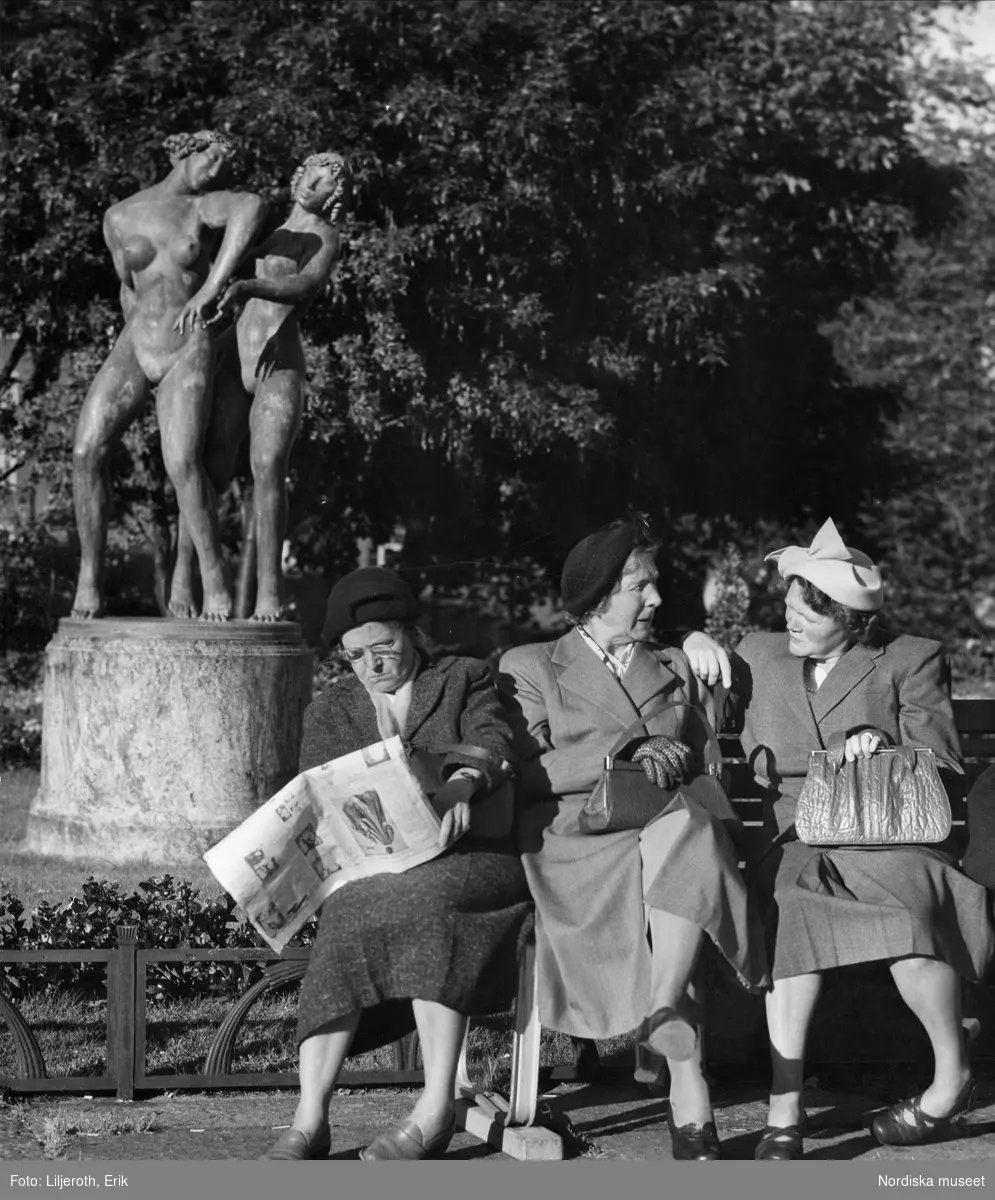 Tre kvinnor sitter på en bänk vid Götaplatsen i Göteborg, två av dem samtalar medan den tredje läser tidningen. I bakgrunden Carl Milles staty Danserskorna.