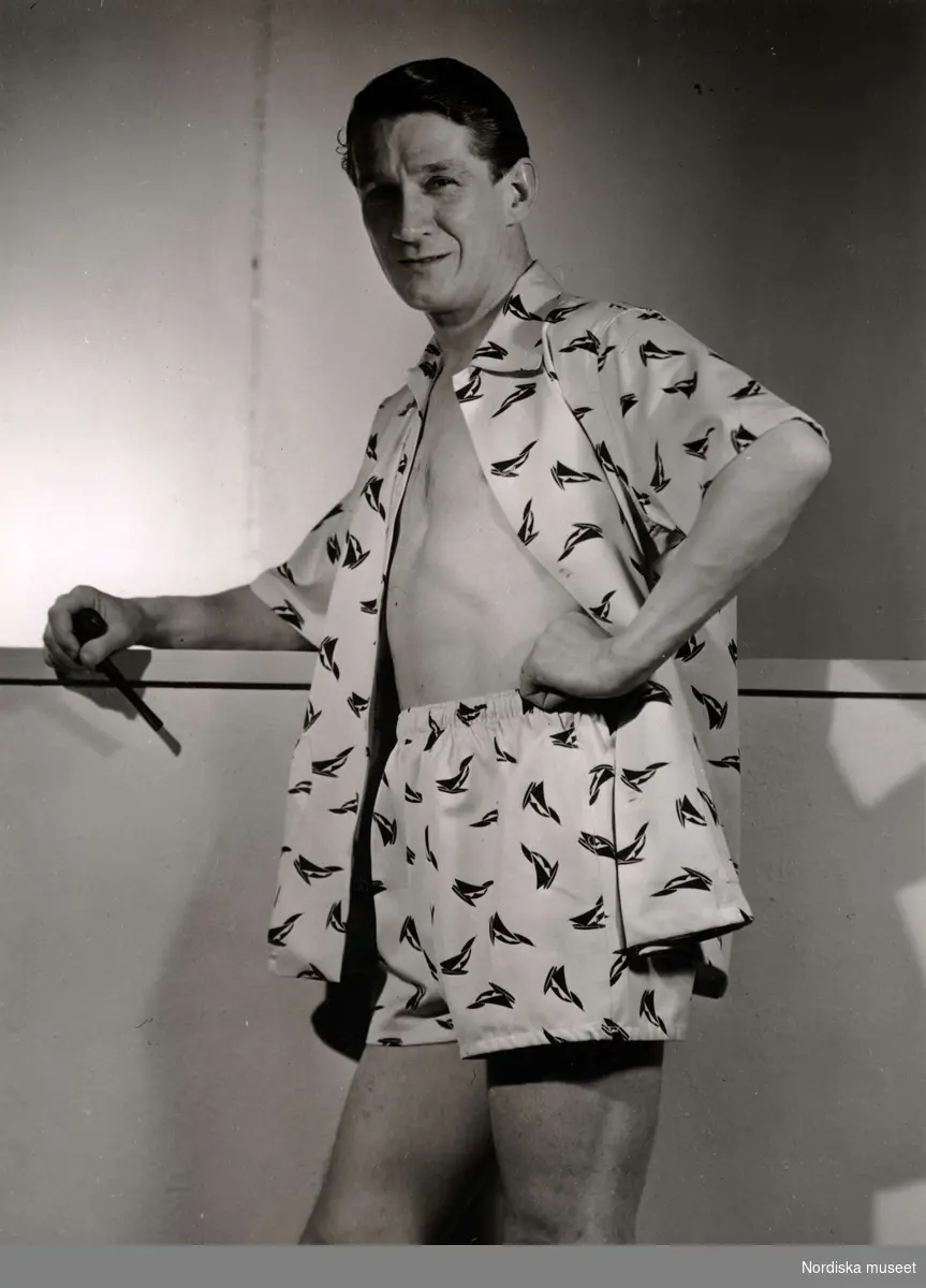 En man visar boxershorts och skjorta (möjligen pyjamas) i samma mönstrade tyg från California - Kläder. I handen håller han en pipa. På varuhuset Nordiska Kompaniet i Stockholm 1947.