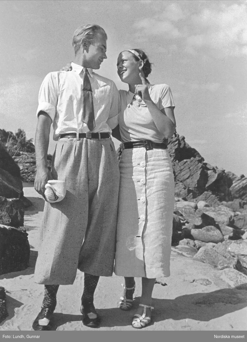 Sommar vid stranden, Sandkaas på Bornholm. Man och kvinna i ledig fritidsklädsel står sida vid sida och håller om varandra, ser varandra djupt i ögonen och ler.