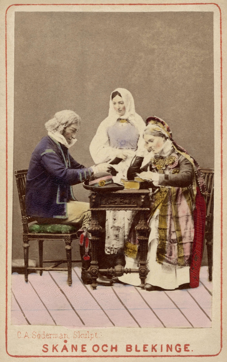 Man och två kvinnor i folkdräkter från Skåne och Blekinge sitter vid bord. Dräktdockor med huvuden skulpterade av C A Söderman, visade på världsutställningen i Paris 1867.