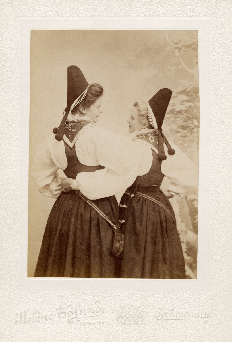 Två kvinnor poserar i folkdräkter, med ryggen vänd mot kameran